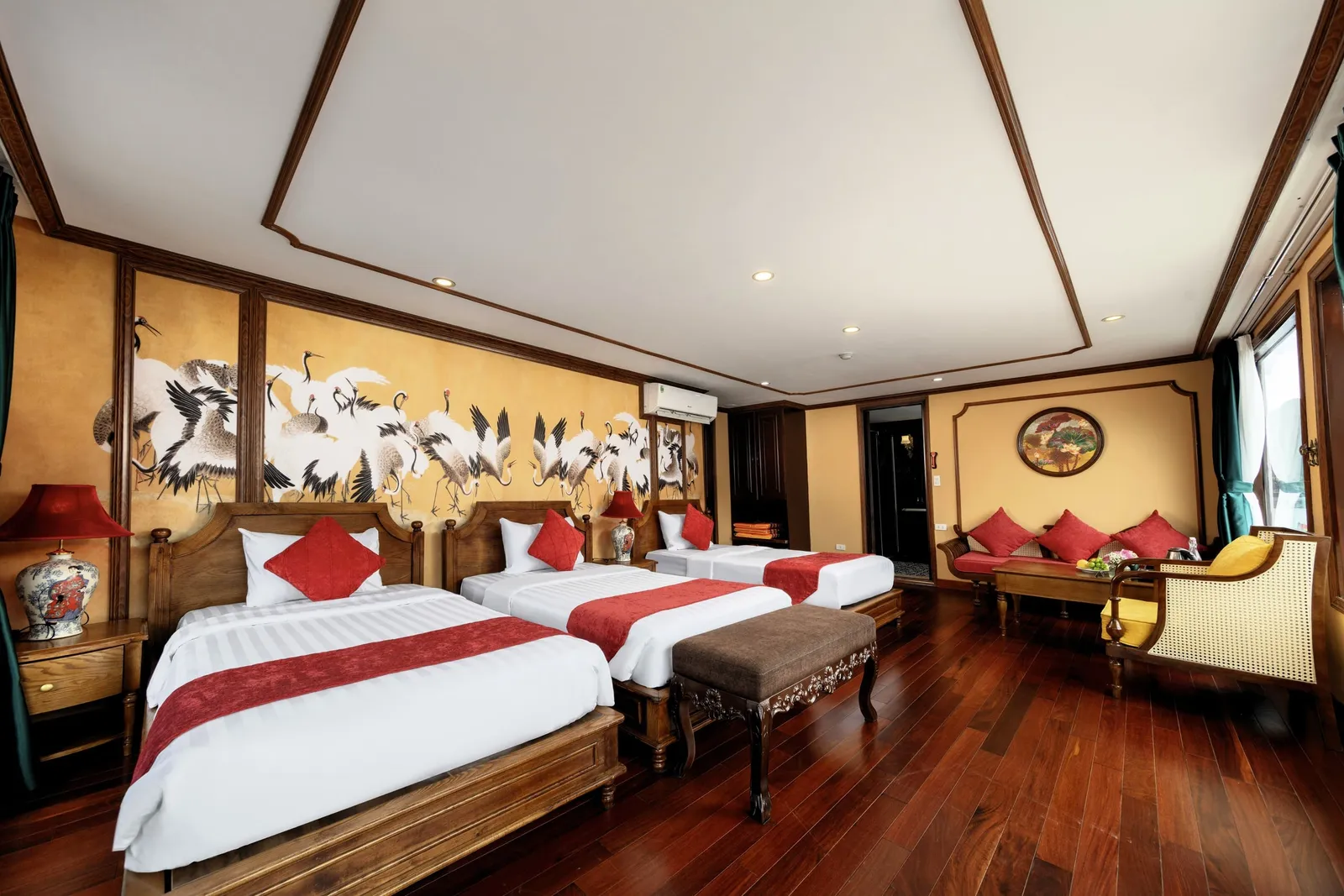 A wood-paneled cabin with three twin beds with white linens and red runners, a painted crane mural on the wall, a sitting area with red cushions, dark hardwood floors, and a window with green curtains.