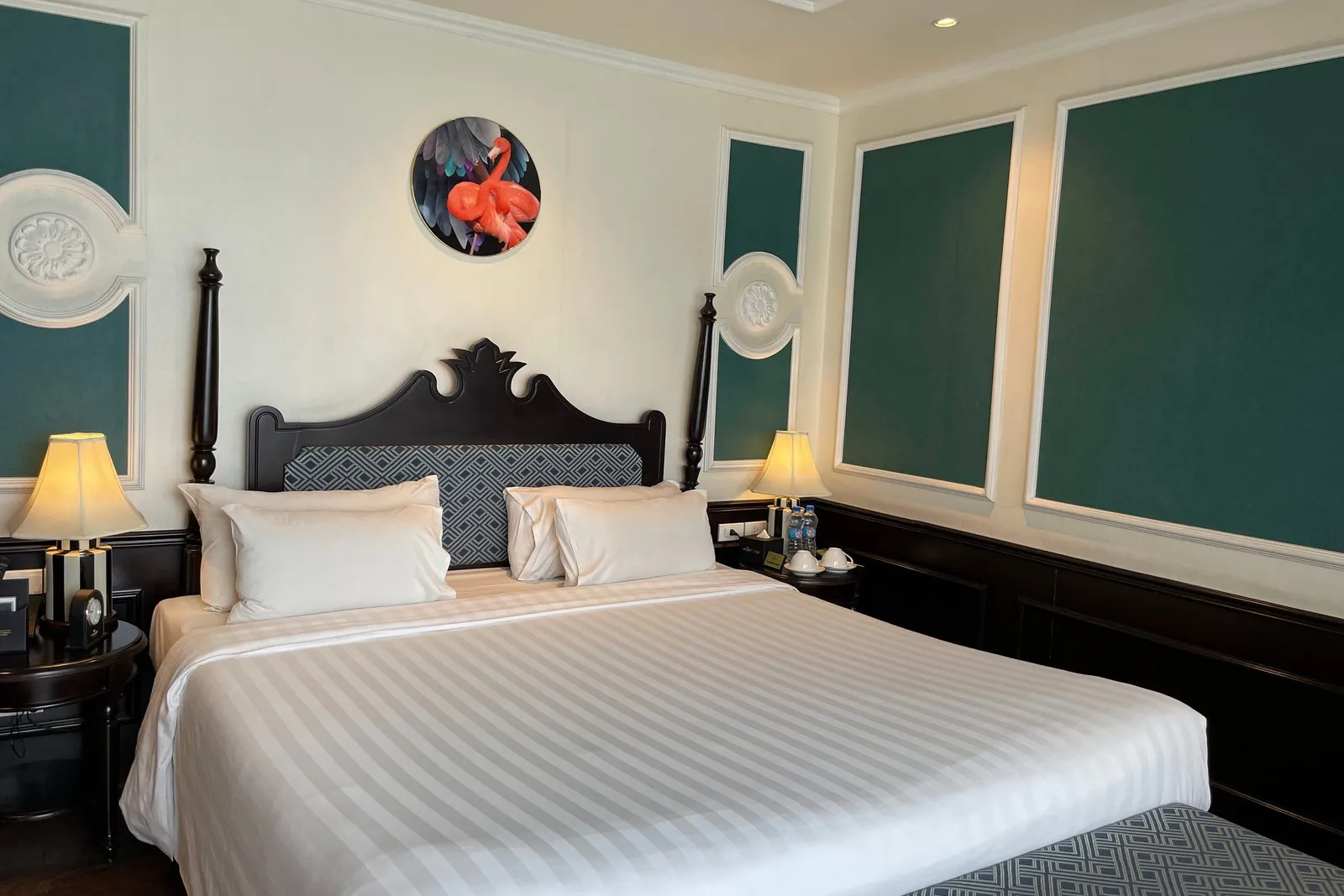 A cabin with a large white-linen bed, dark wood headboard and posts, two lit bedside lamps, teal wall panels with white molding, and a round flamingo artwork above the bed.