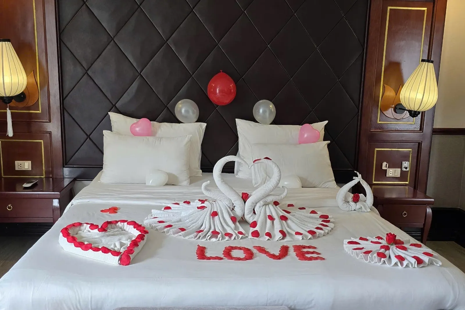 A cabin double bed decorated for a honeymoon with towel-folded swans, a rose-petal heart, the word LOVE spelled in red petals, and balloons on a dark quilted headboard flanked by lantern-style lamps.