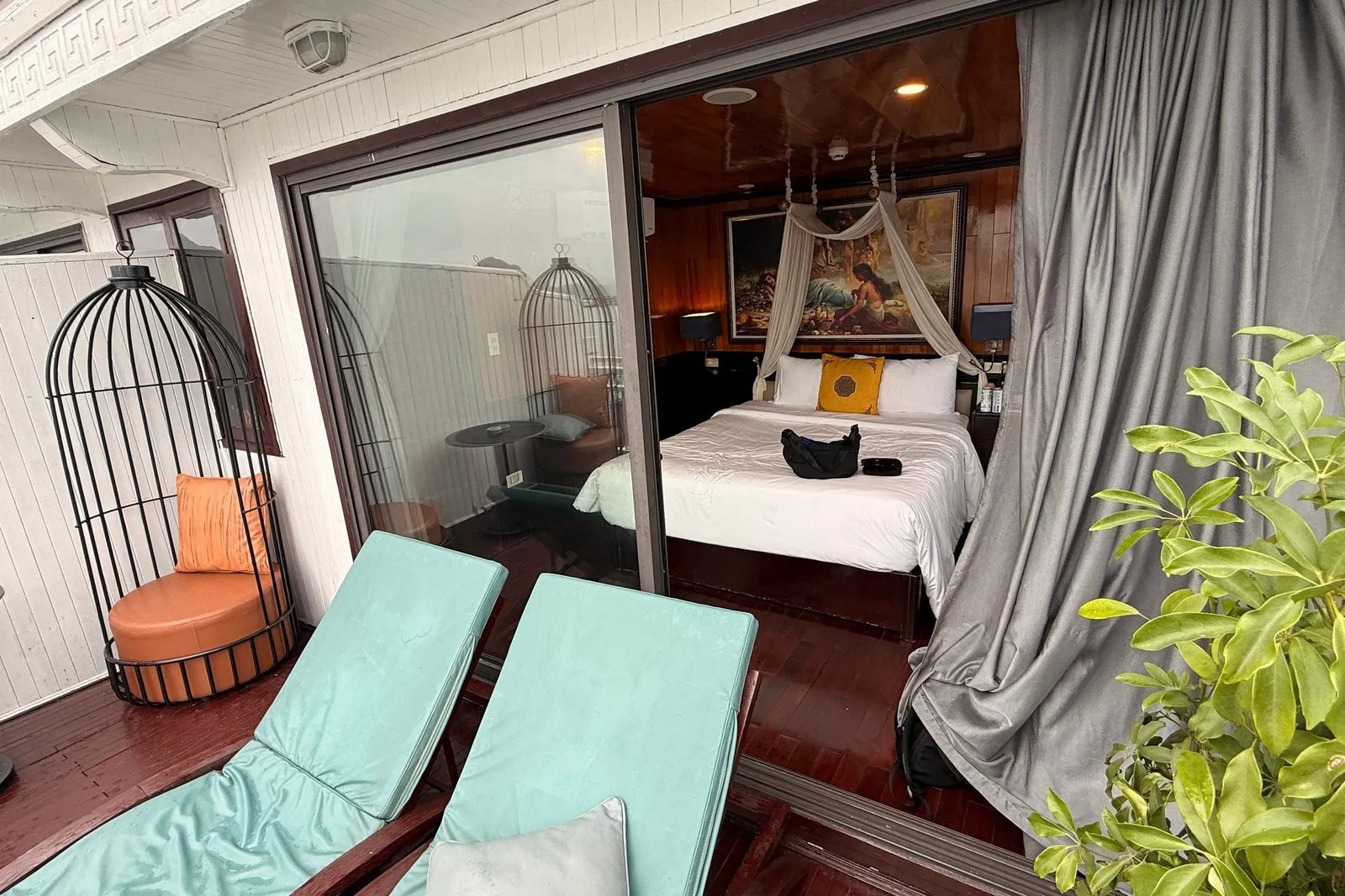A private balcony with two teal sun loungers, a birdcage-style chair and a potted plant, looking through open glass doors into a wood-paneled cabin with a canopy double bed and a framed painting.