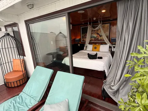 A private balcony with two teal sun loungers, a birdcage-style chair and a potted plant, looking through open glass doors into a wood-paneled cabin with a canopy double bed and a framed painting.