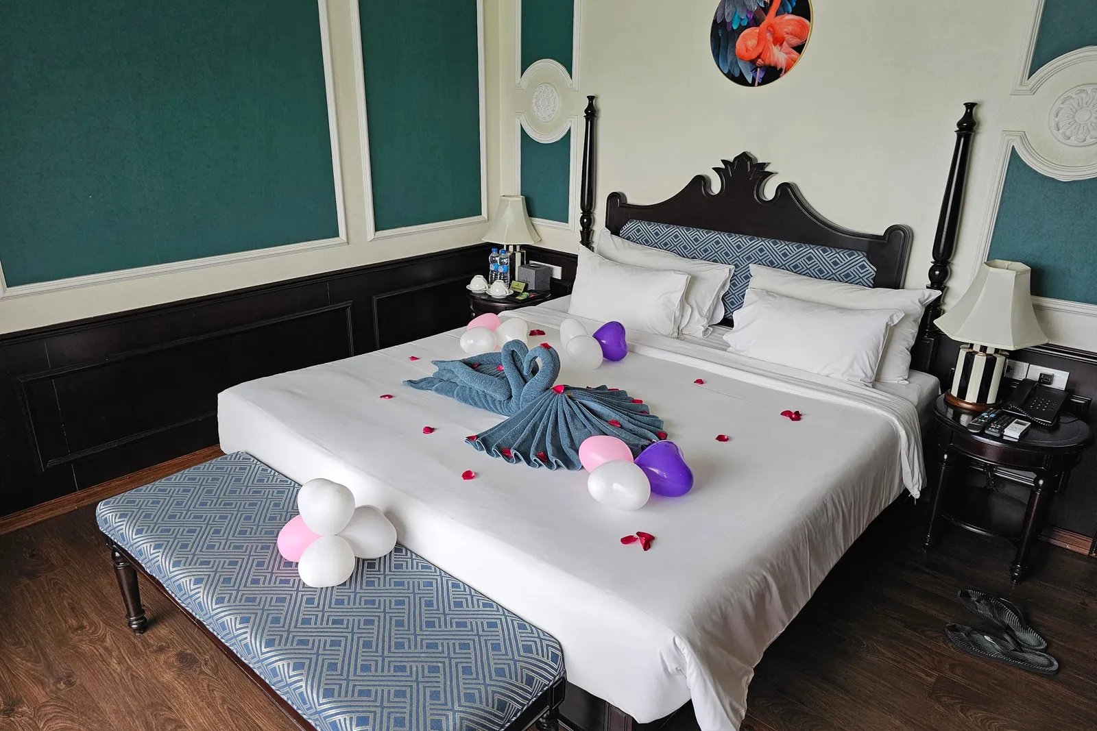 A cabin with a large white bed decorated with a towel swan, heart-shaped balloons and rose petals, a blue patterned bench at the foot, dark wood paneling, teal wall panels, bedside lamps and a flamingo picture on the wall.