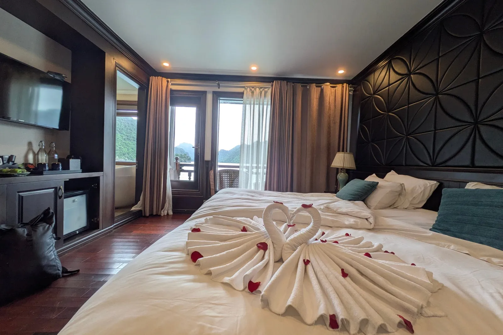 A dark-wood cabin with a large bed topped with towel swans and red rose petals, a wall-mounted TV, minibar, and balcony doors looking out to green karst hills.