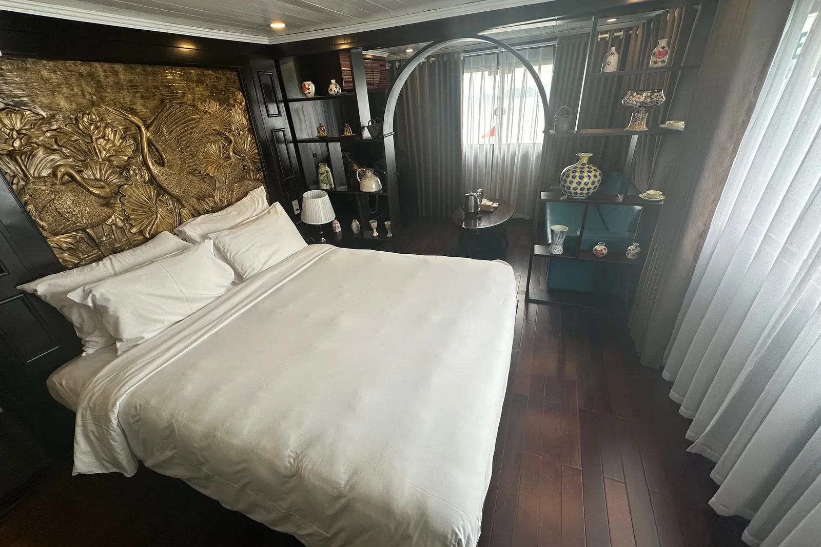 A cruise cabin with a white-linen double bed under a gold relief headboard of cranes and lotus, dark wood floor, shelves of ceramic vases, and sheer curtains over windows.