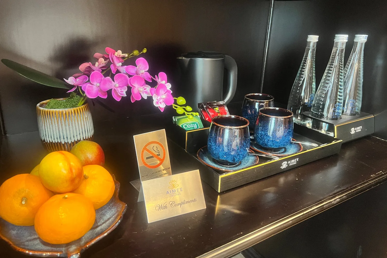 A dark wooden cabin counter holds a plate of oranges, a pink orchid in a pot, blue ceramic cups on a tray, a black kettle, water bottles, and a 'With Compliments' card and no-smoking sign, with Aimee Cruises branding on the card and trays.