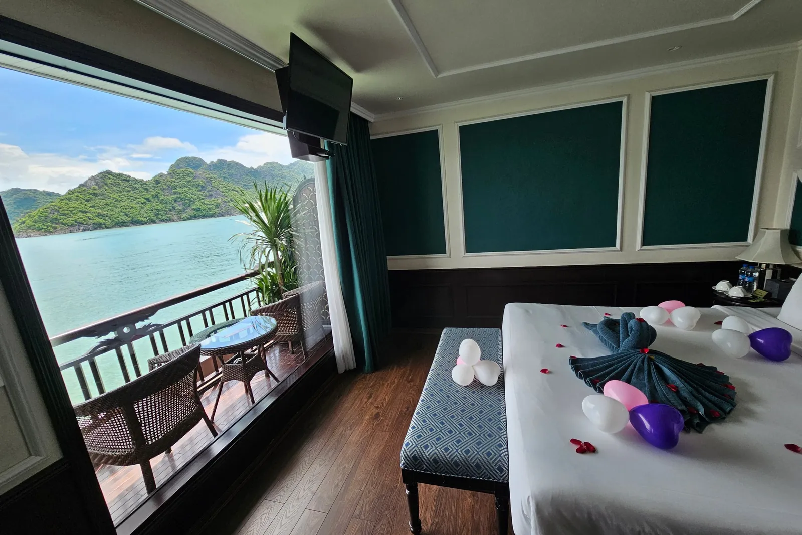 A cruise cabin with a decorated bed (towel folding, balloons, rose petals), a bench, and a wall-mounted TV, with a large window opening onto a private balcony with wicker chairs and a table overlooking turquoise water and green limestone karst islands.