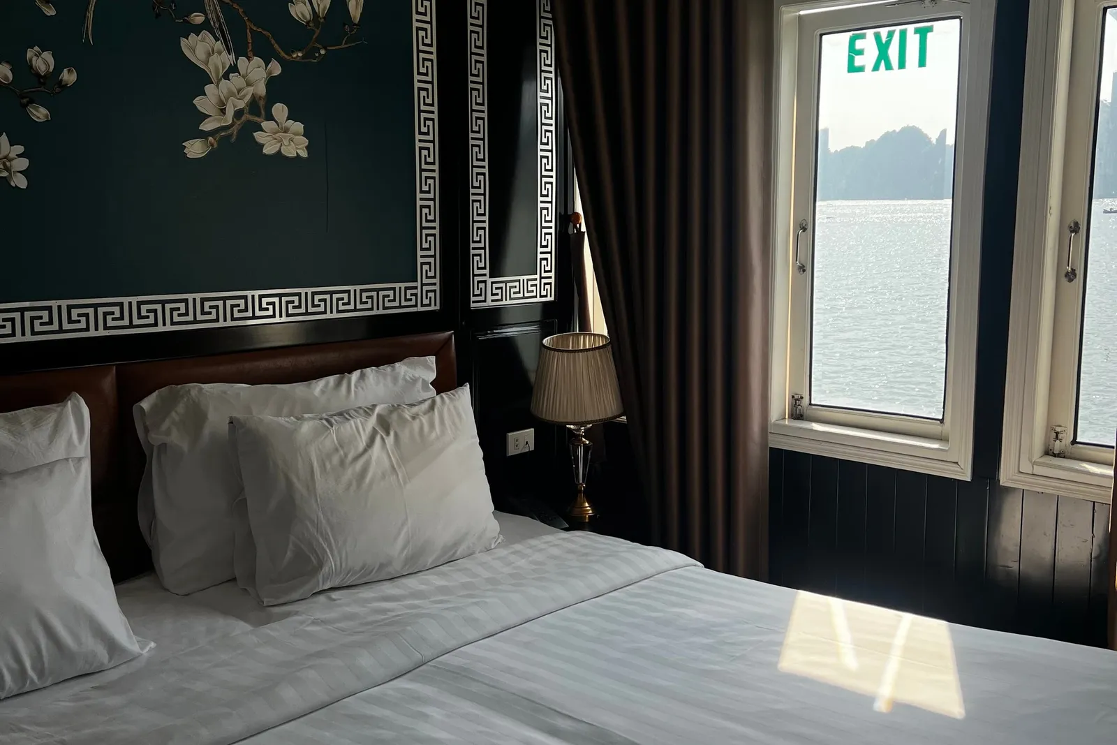 A cruise cabin with a white-linen double bed, dark teal wall panel with magnolia and bird artwork, brown curtains, and a window showing calm water and distant limestone karst islands, with a green EXIT sign visible in the glass.
