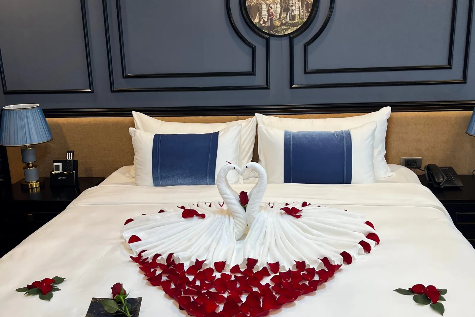 A cabin double bed with white linen and blue velvet-banded pillows, decorated with two towel swans forming a heart and a heart of red rose petals, in front of a navy paneled wall with a round vintage picture.