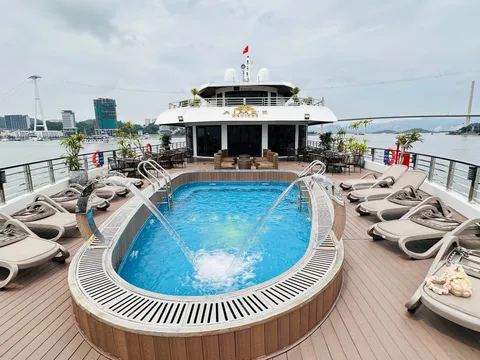 A cruise ship sun deck with an oval plunge pool, loungers and railings, with a branded sign on the upper cabin front and a cable car, bridge and city skyline across the water.