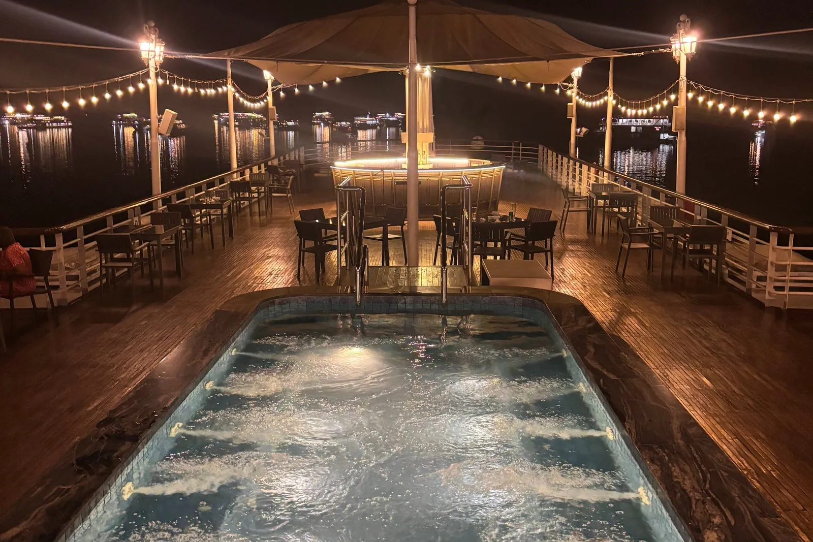 A night view along a boat's upper deck with a bubbling jacuzzi pool in the foreground, tables and chairs, a lit canopy bar, string lights and distant lit boats on dark water.