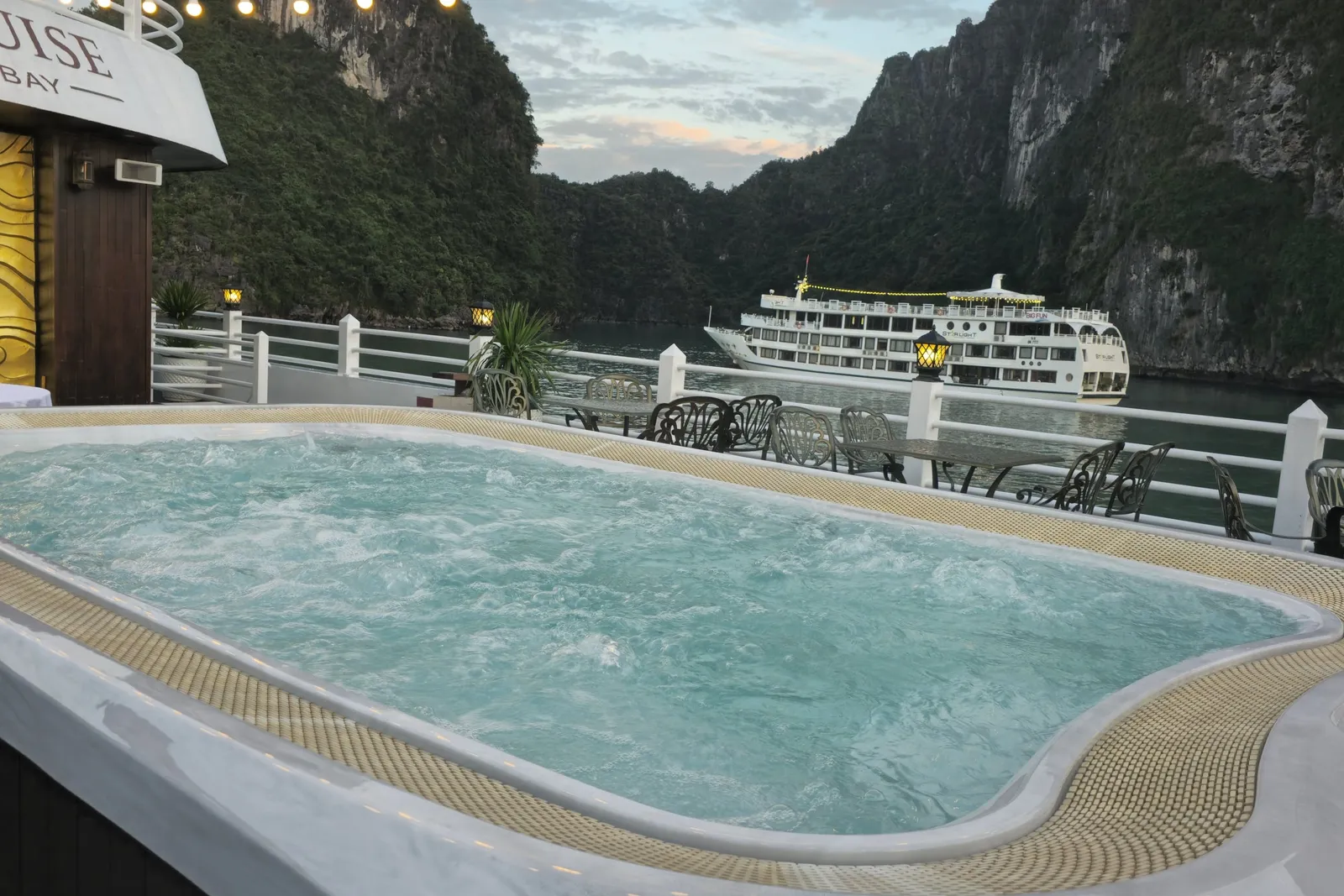 A bubbling jacuzzi on a cruise ship's sun deck with string lights and outdoor tables, looking toward limestone karst cliffs and another white cruise ship on the water at dusk, with partial lettering ending in 'UISE' and 'BAY' on the wall of the superstructure.
