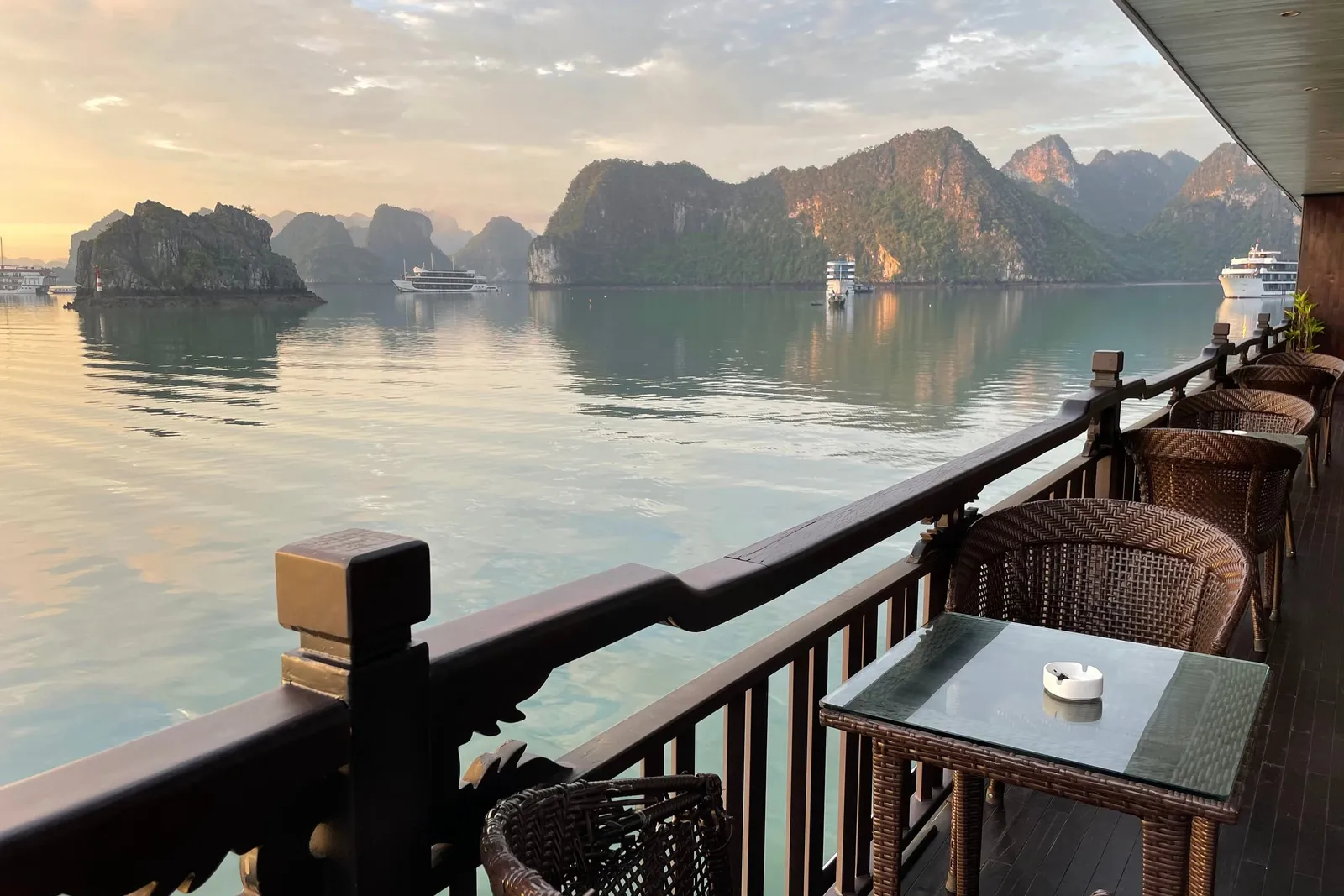 A wooden railing and wicker chairs with a glass-topped table on a boat deck, looking out over calm water toward limestone karst islands and distant cruise boats at sunrise.