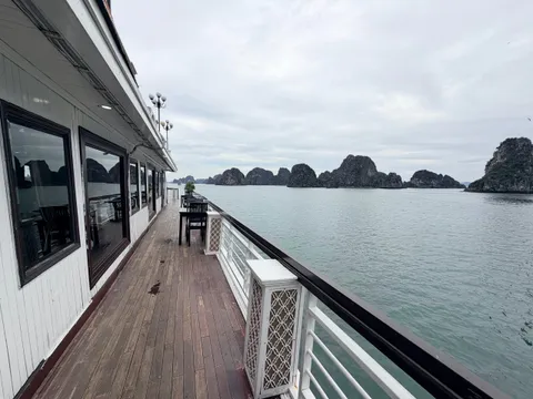 A wooden side deck of a boat with a white railing, dark chairs and a table, and cabin windows, looking out over calm green water toward limestone karst islands under an overcast sky.