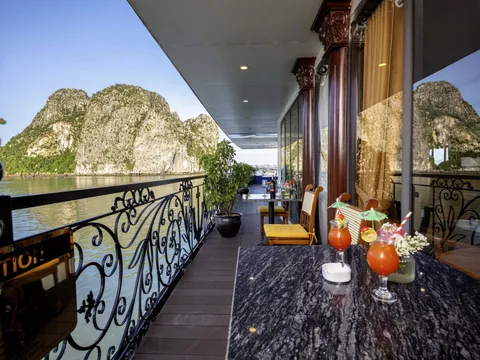 A covered wooden boat deck with wrought-iron railing, a dark marble table holding two orange cocktails and a flower vase, yellow-cushioned chairs, potted plants, and limestone karst islands across calm green water, with a partly visible gold-lettered sign in the lower left corner.