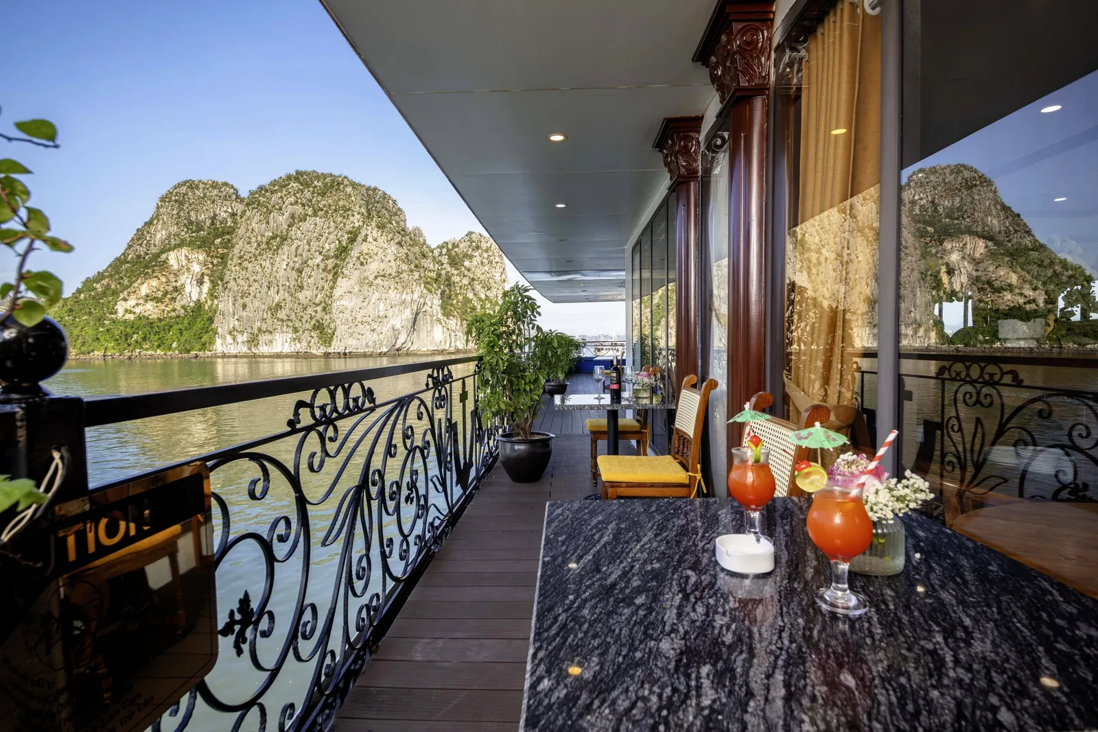 A covered wooden boat deck with wrought-iron railing, a dark marble table holding two orange cocktails and a flower vase, yellow-cushioned chairs, potted plants, and limestone karst islands across calm green water, with a partly visible gold-lettered sign in the lower left corner.