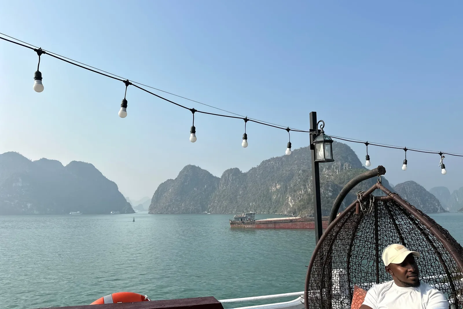 A man in a white cap and t-shirt sits in a hanging wicker egg chair on a boat deck under string lights, facing a calm green bay with limestone karst islands and a cargo barge in the hazy distance.