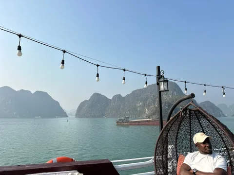 A man in a white cap and t-shirt sits in a hanging wicker egg chair on a boat deck under string lights, facing a calm green bay with limestone karst islands and a cargo barge in the hazy distance.