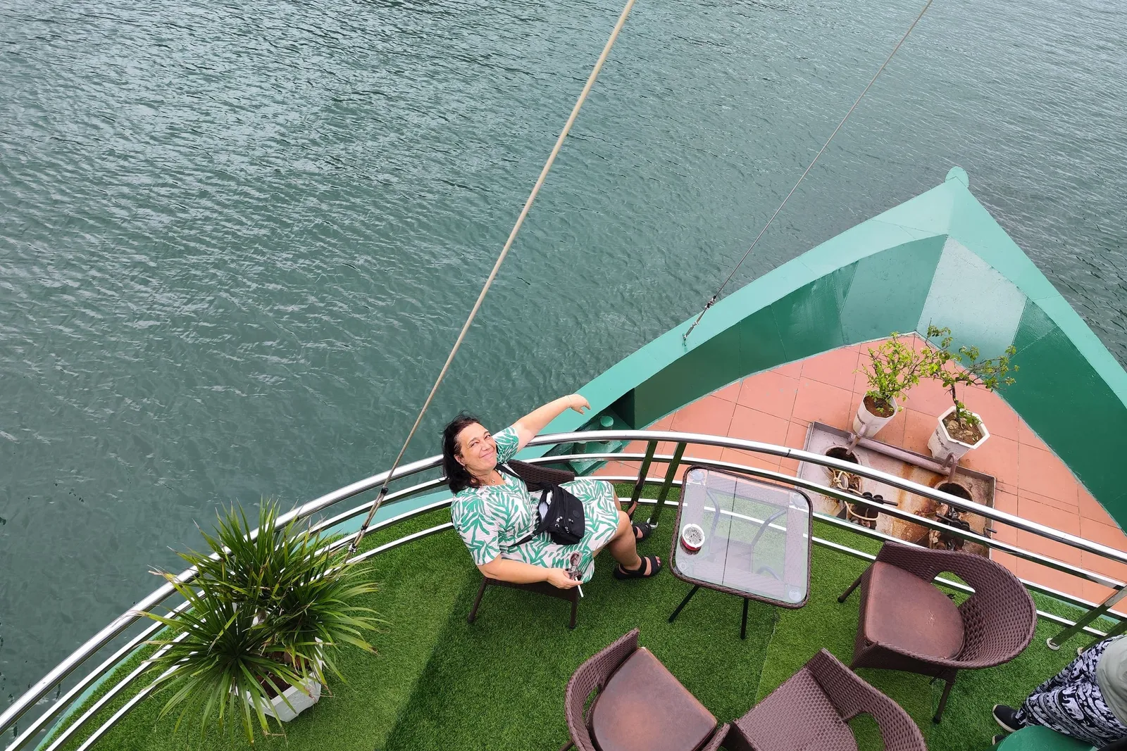 A high-angle view down onto a boat's green bow deck with artificial turf, wicker chairs, a glass table and potted plants, where a smiling woman in a green patterned dress sits waving beside the railing above calm green water.