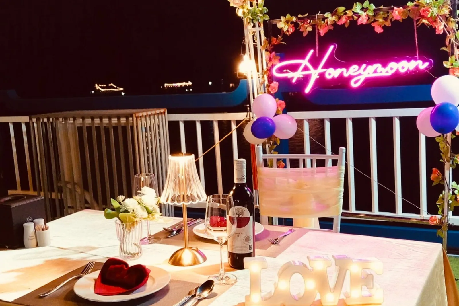 A night-time romantic dinner table on a boat deck with a pink neon 'Honeymoon' sign, a lit 'LOVE' marquee, balloons, flowers, a wine bottle and a heart-shaped napkin, with distant lights across the water.