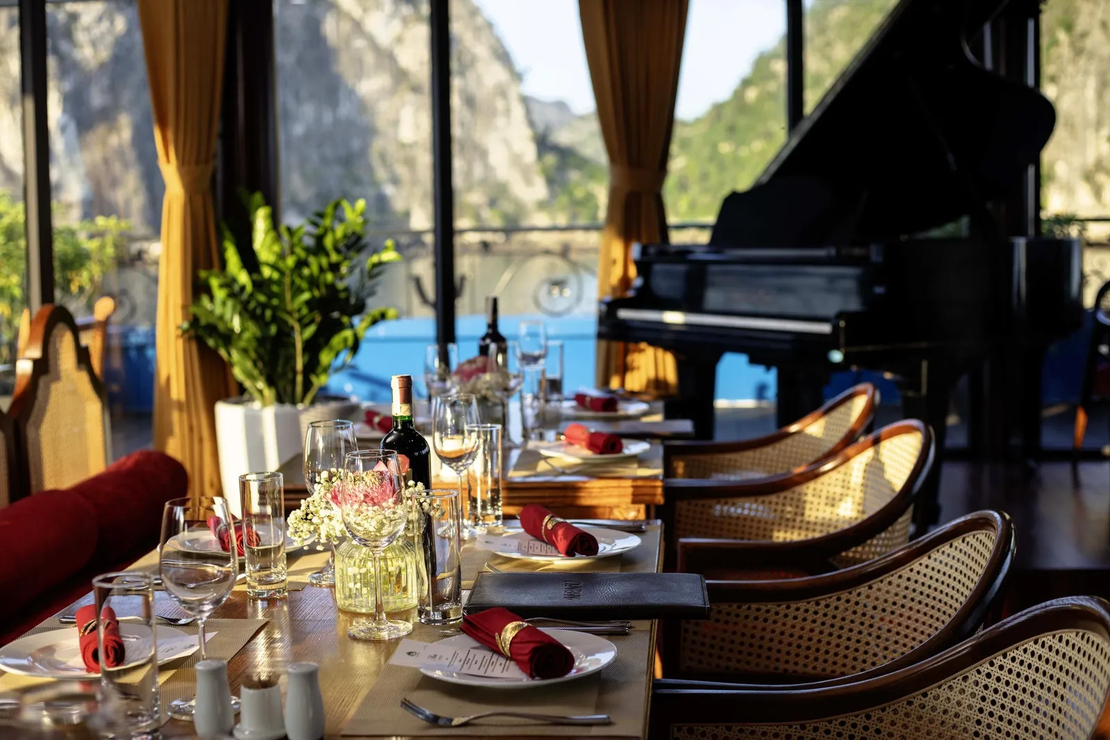 A set dining table with wine glasses, red napkins and rattan chairs sits beside a grand piano in a lounge with large windows showing green limestone karst cliffs and blue water outside.