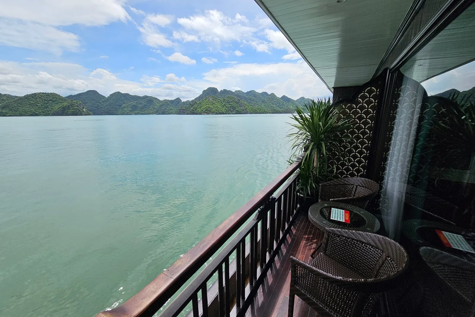 A private balcony on a boat with wicker chairs, a small table and a potted plant, overlooking turquoise water and green karst islands under a partly cloudy sky.