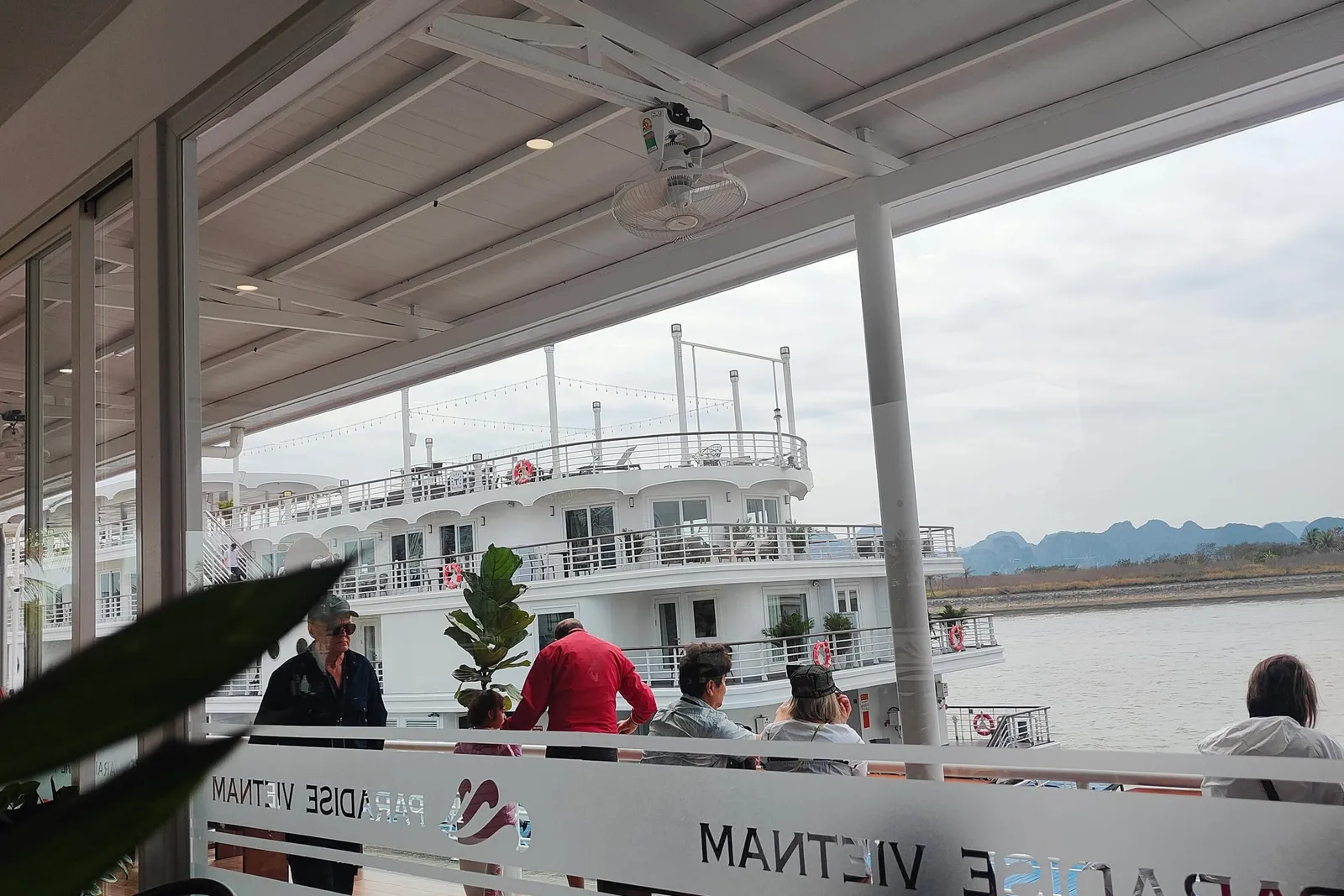 View from inside a covered lounge through a glass panel etched with a mirrored 'Paradise Vietnam' logo, showing passengers on an outdoor deck, a multi-level white cruise ship alongside, and water with distant limestone hills.
