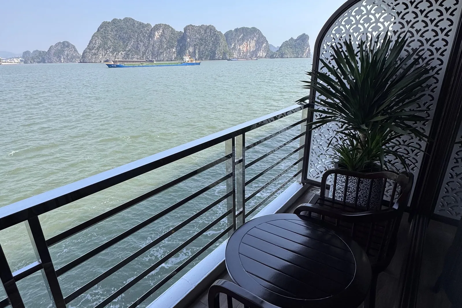 A private cabin balcony with a dark wooden table, two chairs, a potted plant and a decorative lattice screen, overlooking green water, limestone karst islands and a cargo ship.