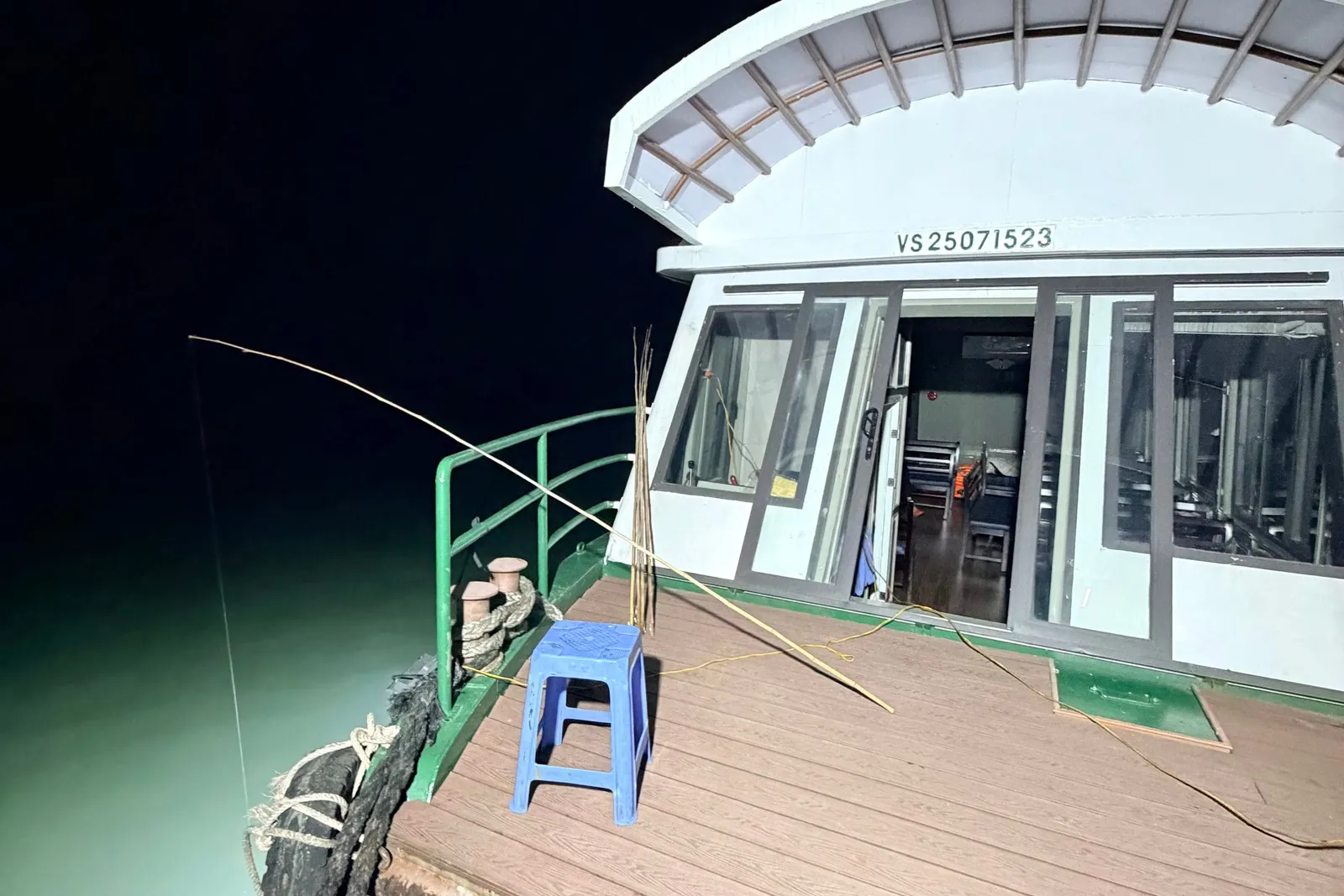 A night shot of a boat's wooden-look bow deck with a blue plastic stool, a bamboo fishing rod over the side, mooring bollards, rope and tires as fenders, and a white cabin with sliding doors showing the registration number VS 25071523 above it, over dark green-lit water.