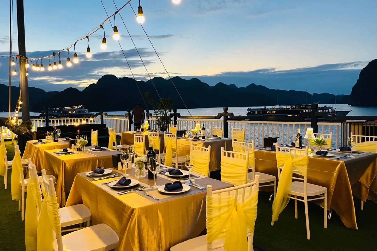 Dusk view from an upper deck set with tables in gold cloths and white chairs with yellow sashes, string lights overhead, and limestone karst islands and other boats on the water beyond the railing.