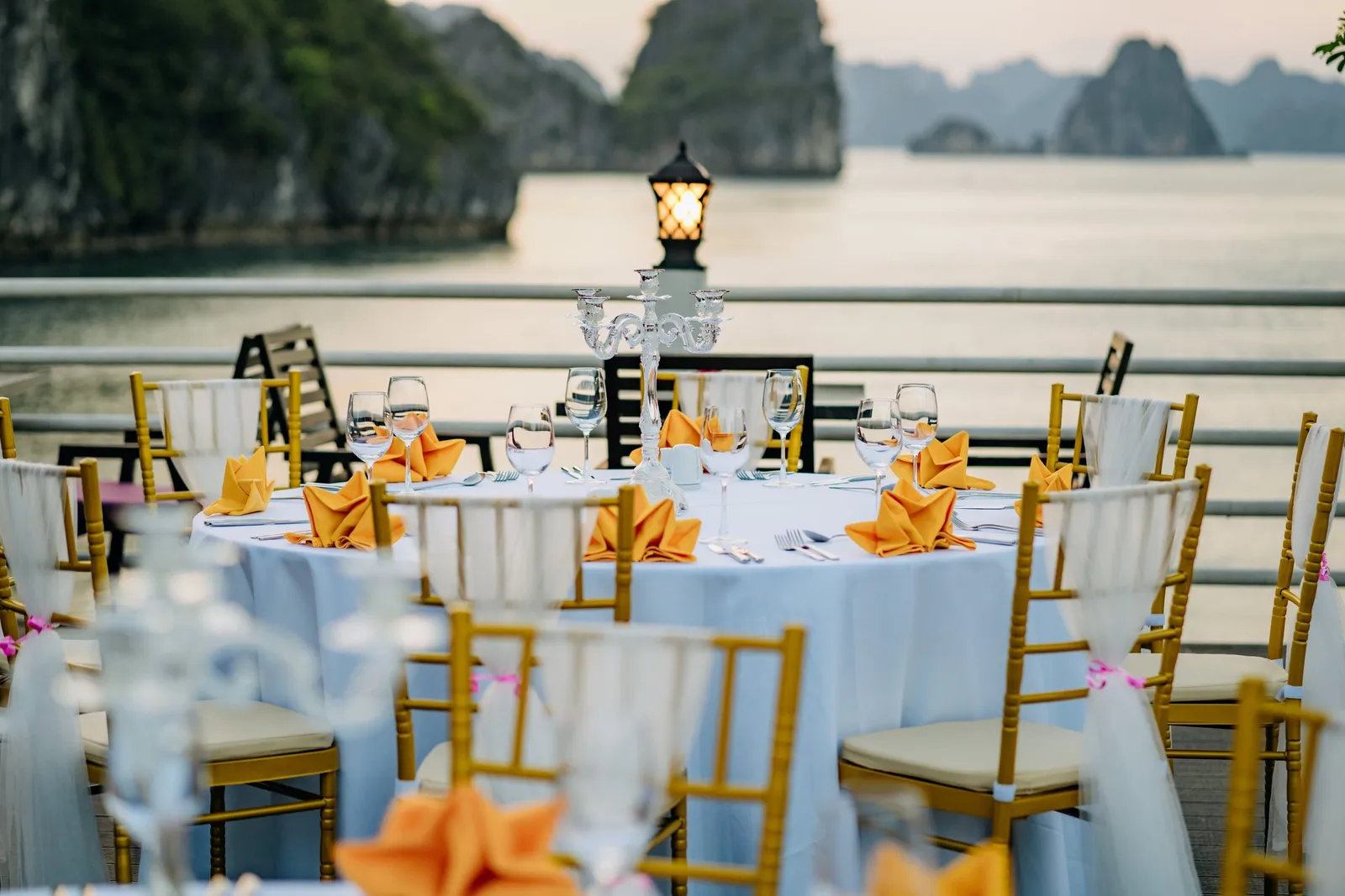 A round table with white cloth, orange napkins, wine glasses and a candelabra, surrounded by gold chairs with white sashes on an open deck overlooking calm water and limestone karst islands at dusk.
