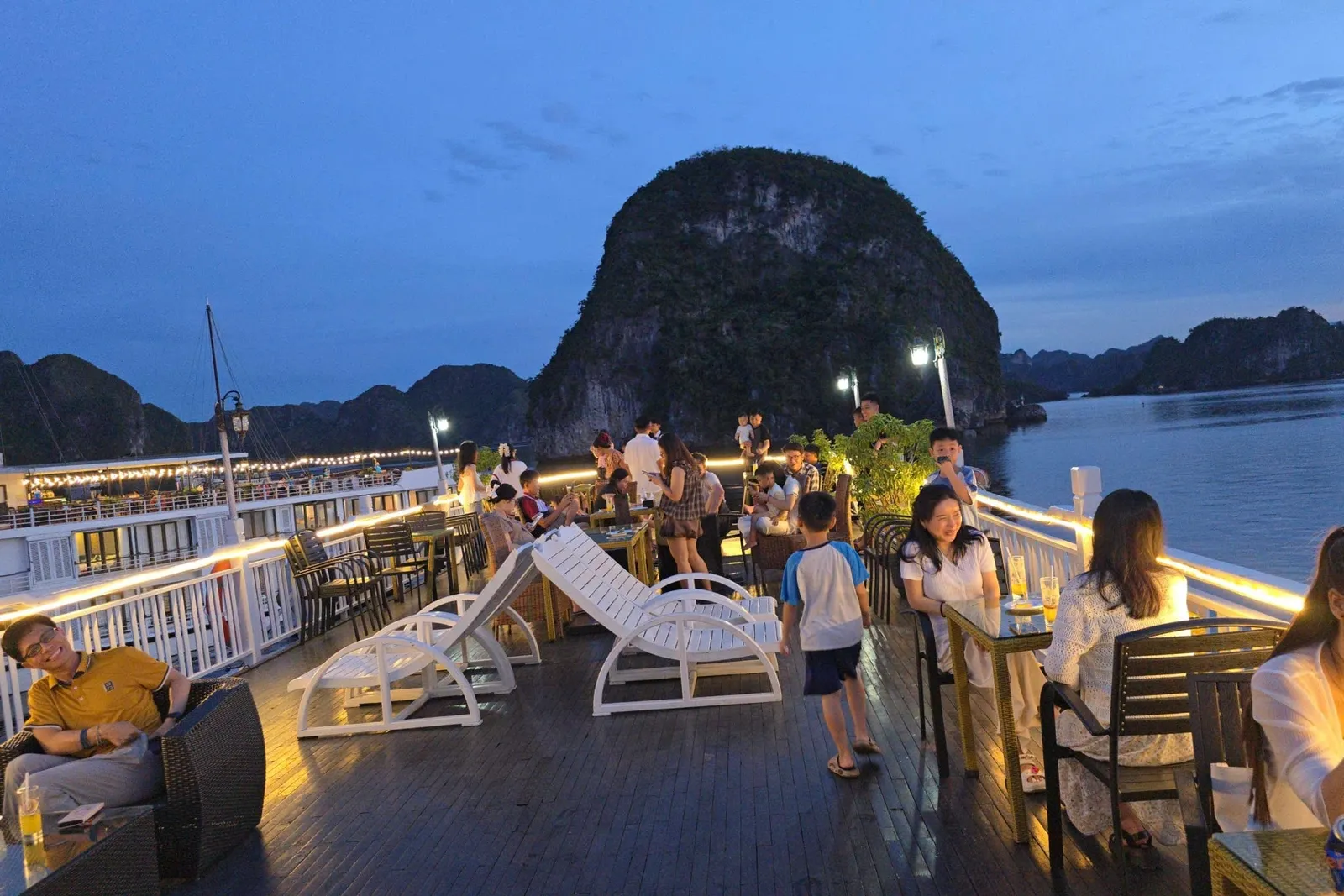 At dusk, guests sit at tables and loungers on the lit upper deck of a cruise boat, with another boat alongside and a large forested limestone karst rising from the water behind.