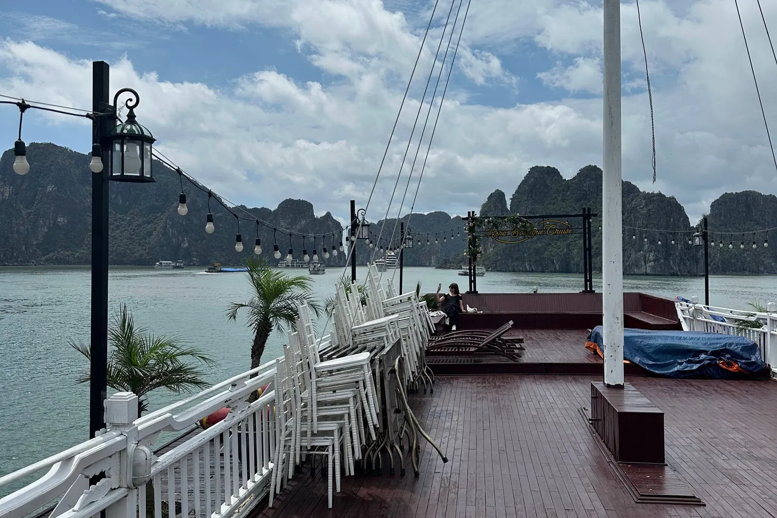 A wet wooden upper deck with white railings, stacked white chairs, string lights, a mast, and a sign reading a cruise name at the far end, with limestone karst islands and a cloudy sky beyond calm green water.