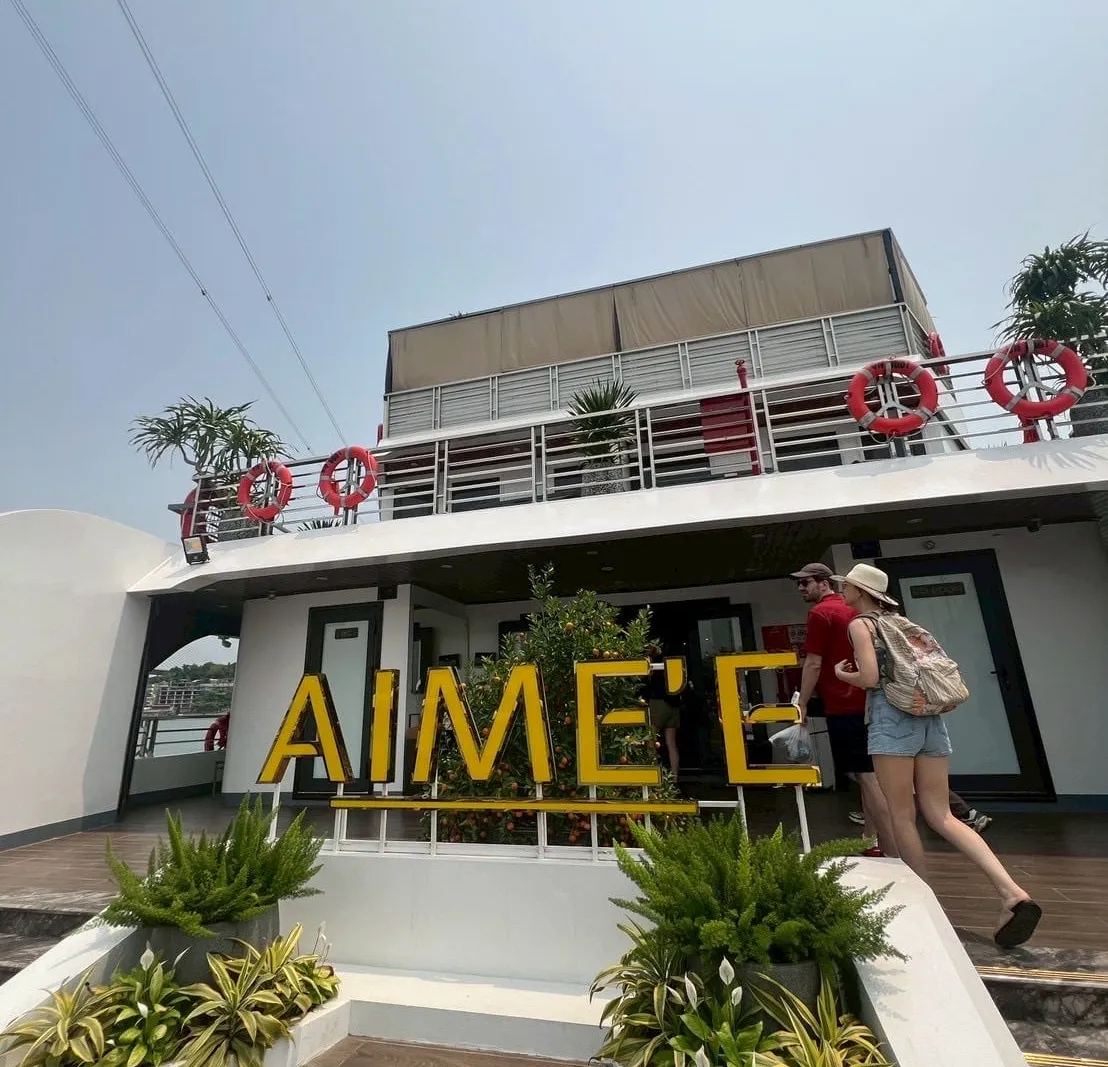 Upward view from the deck of a white boat with a large yellow 'AIME'E' sign among planters, red life rings on the upper railing, and two people in the background walking toward the entrance.