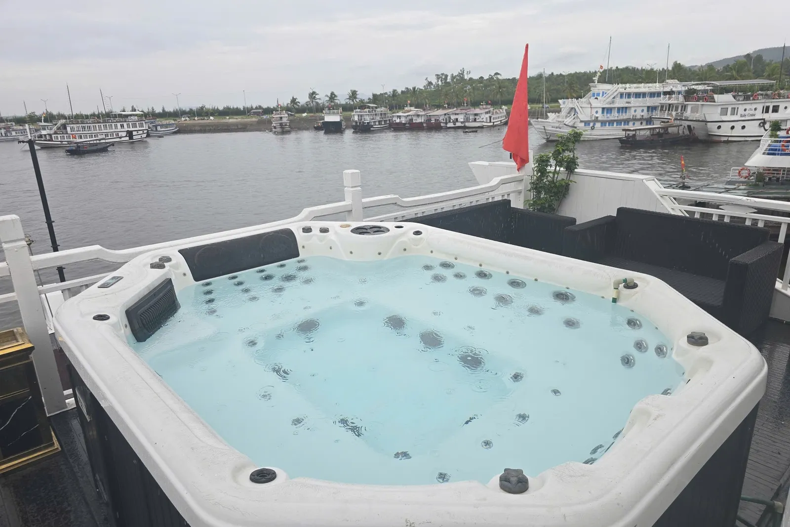 A white hot tub with jets filled with water sits on a boat's upper deck beside black wicker sofas, a red flag and a white railing, overlooking a harbor with many moored white cruise boats under an overcast sky.