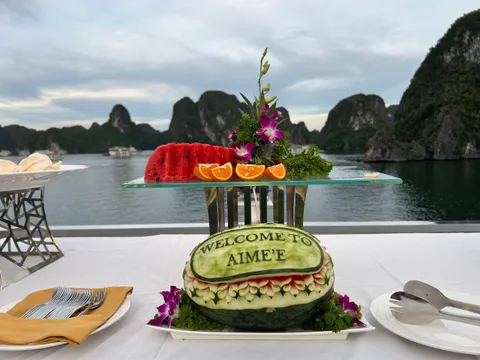 A carved watermelon reading 'Welcome to Aime'e' sits on a white-clothed deck table with sliced watermelon, orange segments, orchids and cutlery, in front of calm water and limestone karst islands at dusk.