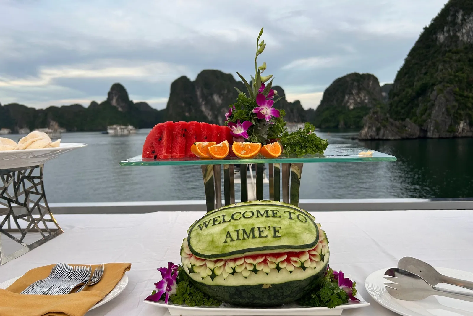 A carved watermelon reading 'Welcome to Aime'e' sits on a white-clothed deck table with sliced watermelon, orange segments, orchids and cutlery, in front of calm water and limestone karst islands at dusk.
