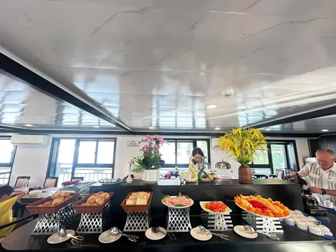 A breakfast buffet counter in an indoor dining room with bread baskets, sliced ham, tomatoes, fruit platter, flower vases and serving tongs, with windows and a railing behind, an older man pouring from a kettle on the right, and a woman standing behind the counter.