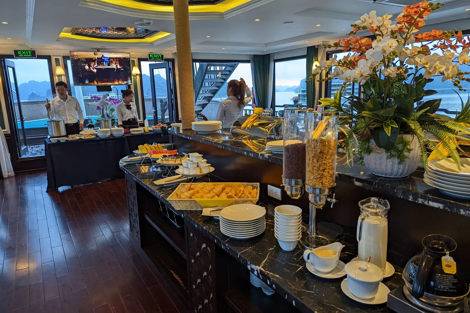 A breakfast buffet counter with plates, cereal dispensers, pastries, fruit and an orchid arrangement inside a boat dining room, with staff in the background and limestone karsts visible through the windows.