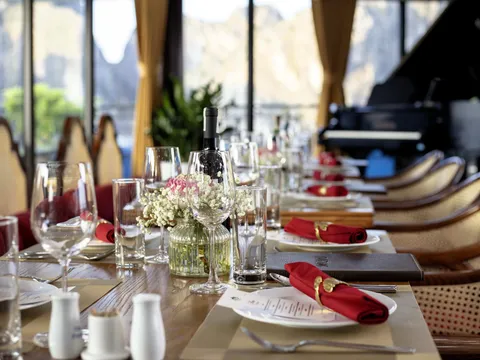 A long dining table set with red napkins, glasses, a flower vase and a menu folder, with rattan chairs, a grand piano and windows showing misty limestone karst hills behind.