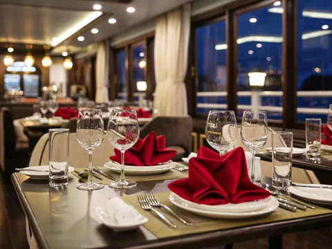 A dining room at night with tables set with red folded napkins, wine glasses, tumblers and cutlery, with large windows showing a railing and dark water outside.