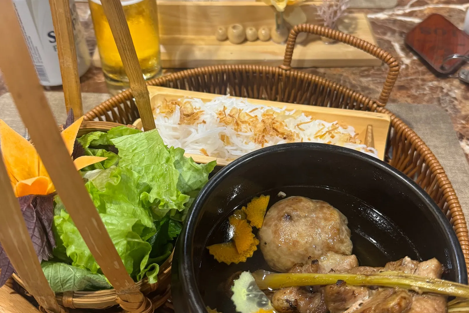 A basket tray holding a black bowl of grilled pork skewer and meatball in broth, rice noodles and fresh lettuce, with a printed set menu card, a beer glass and cans on a marble table.