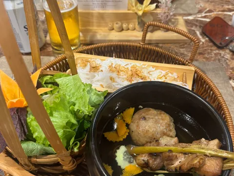 A basket tray holding a black bowl of grilled pork skewer and meatball in broth, rice noodles and fresh lettuce, with a printed set menu card, a beer glass and cans on a marble table.