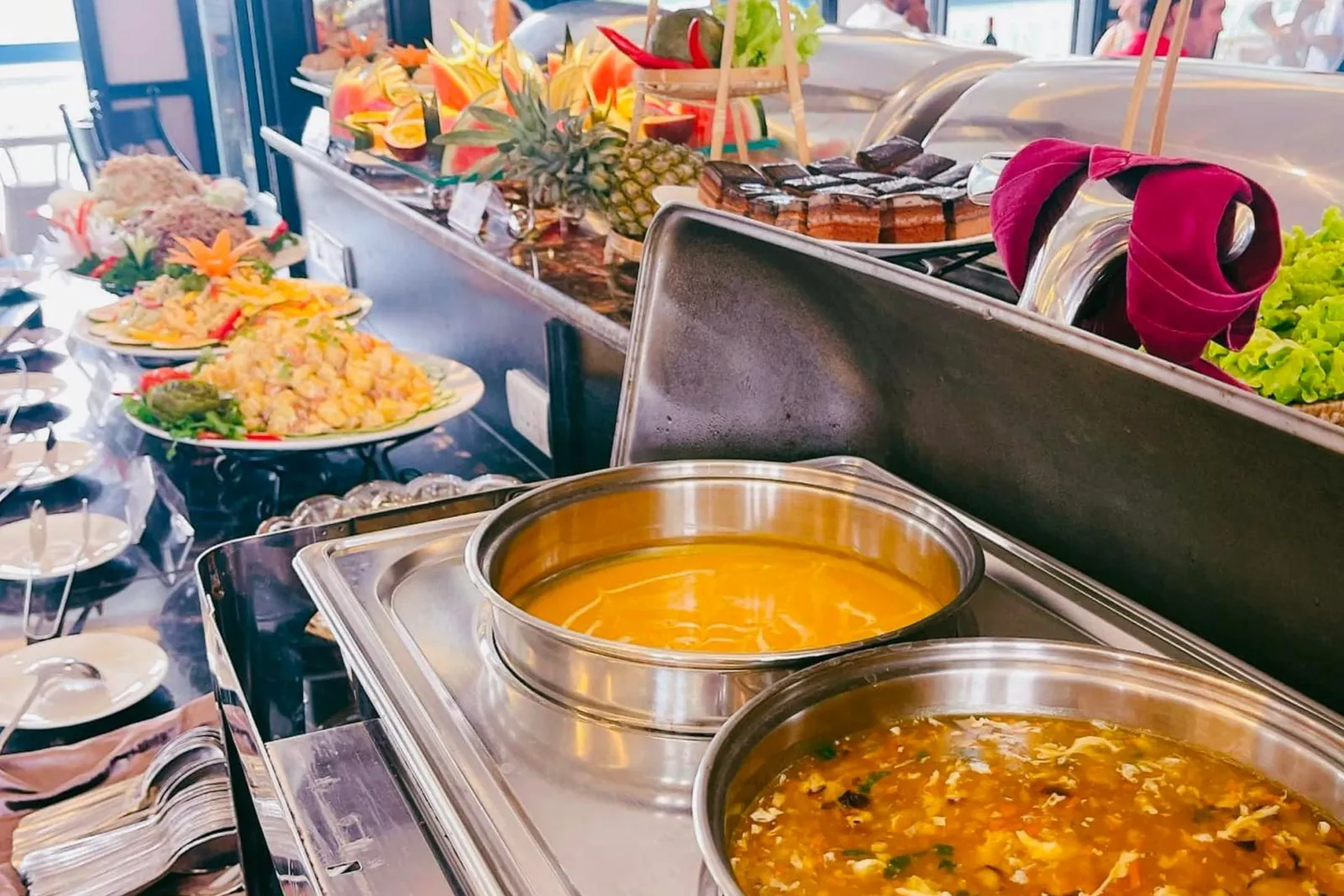 A buffet line with chafing dishes of orange soups, plated salads, carved fruit, a pineapple, chocolate cakes and stacked plates and cutlery, with bright windows in the background.