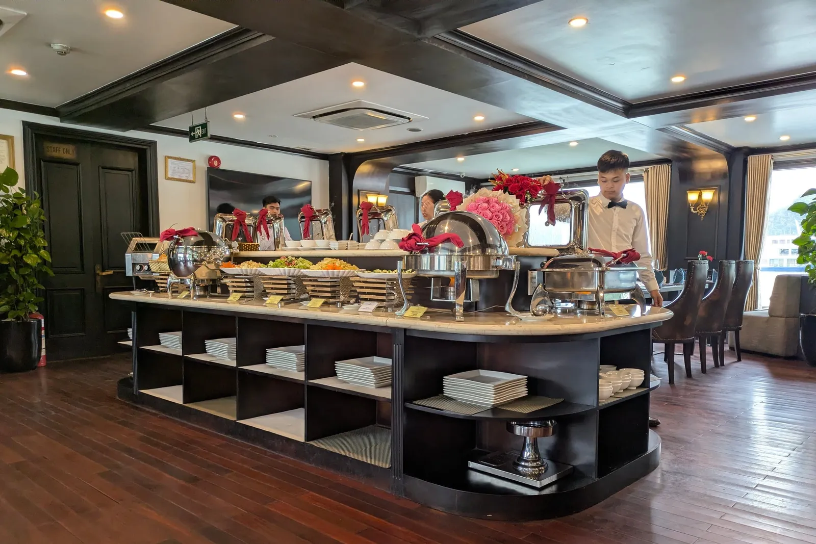 An indoor buffet island with chafing dishes, stacked plates and flower arrangements in a dark-wood dining room, with a waiter in a white shirt and bow tie standing behind it and a window at right.