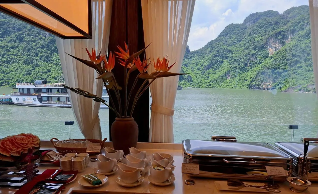 A buffet counter with chafing dishes, cream pitchers, cups and a vase of bird-of-paradise flowers sits in front of a window framing green karst hills, calm green water and another cruise boat.