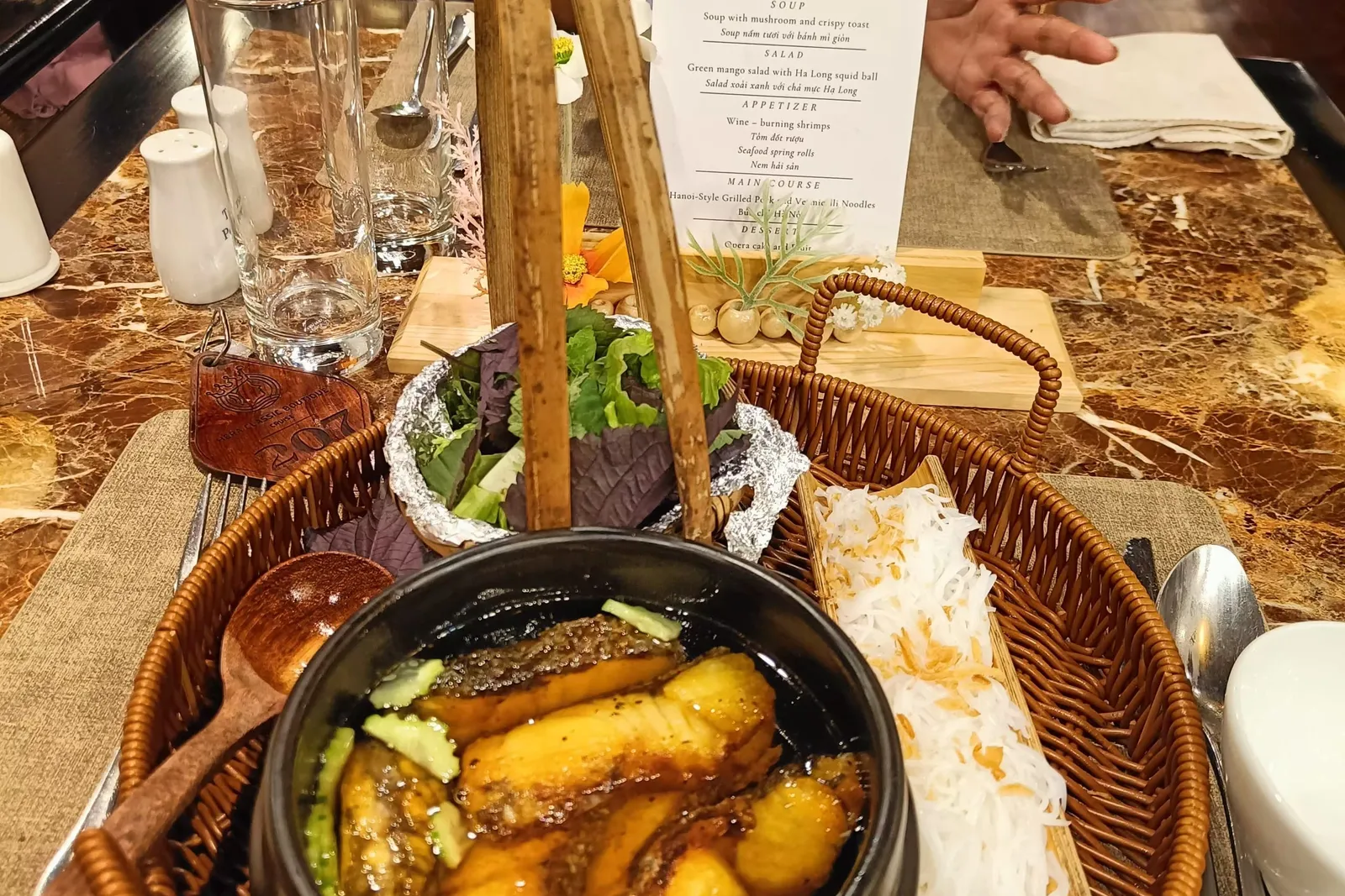 A dining table holds a black clay pot of braised fish in a wicker basket with rice noodles, fresh herbs, a printed set menu card with a logo and Ha Long dishes, and a leather room key tag marked 207, with a seated person gesturing behind it.