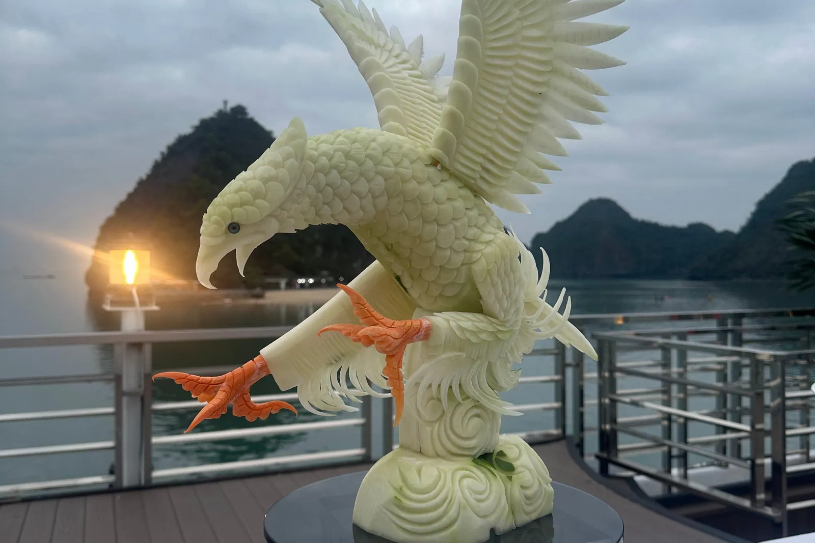 A carved pale-green eagle sculpture with orange talons stands on a glass-topped stand on a white-draped table, on a wooden deck with railings, with limestone islands and a glowing lantern at dusk behind it.