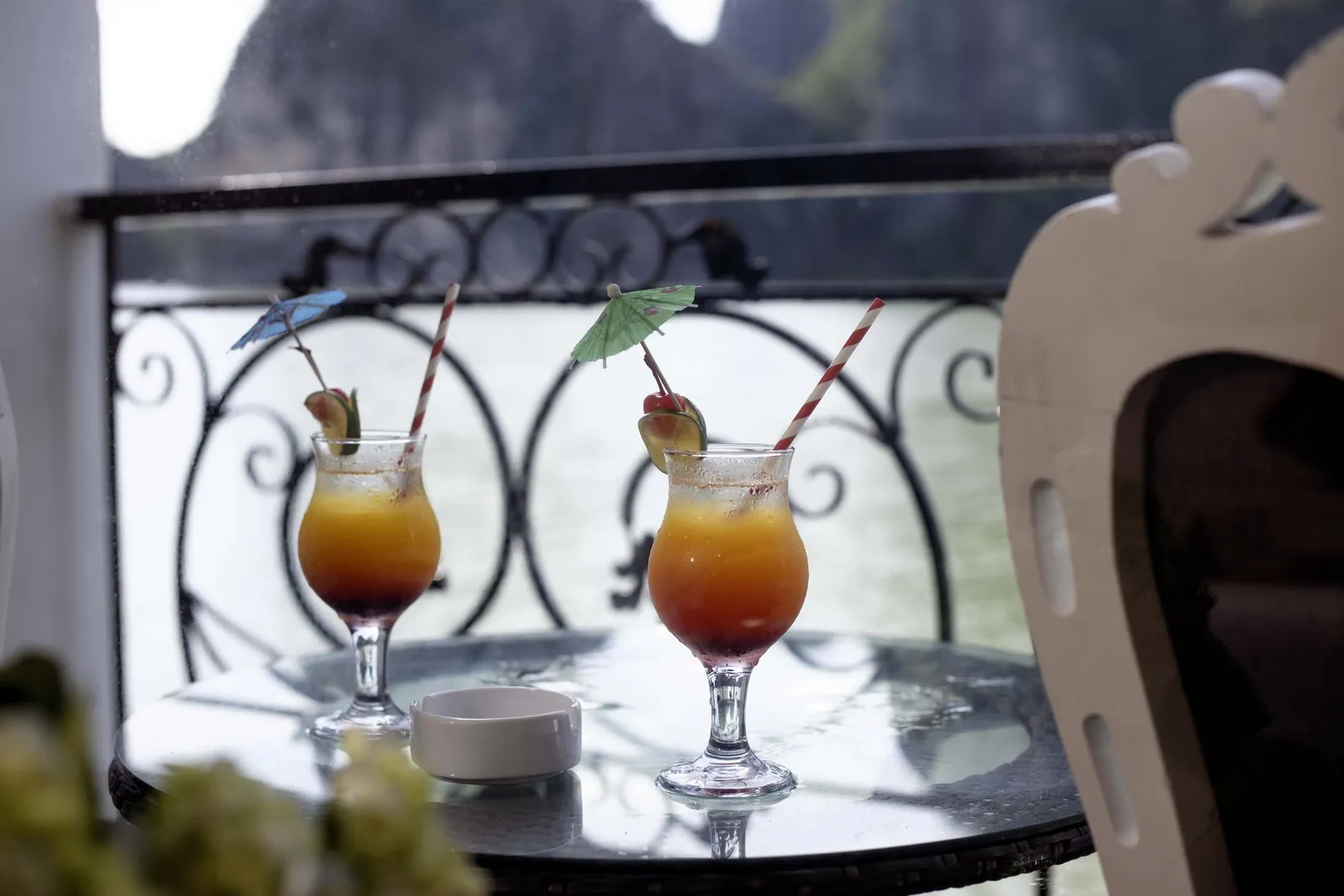 Two orange-and-red cocktails with paper umbrellas, striped straws and fruit garnishes sit on a glass table beside an ashtray, in front of an ornate wrought-iron balcony railing with water and limestone karst cliffs behind.