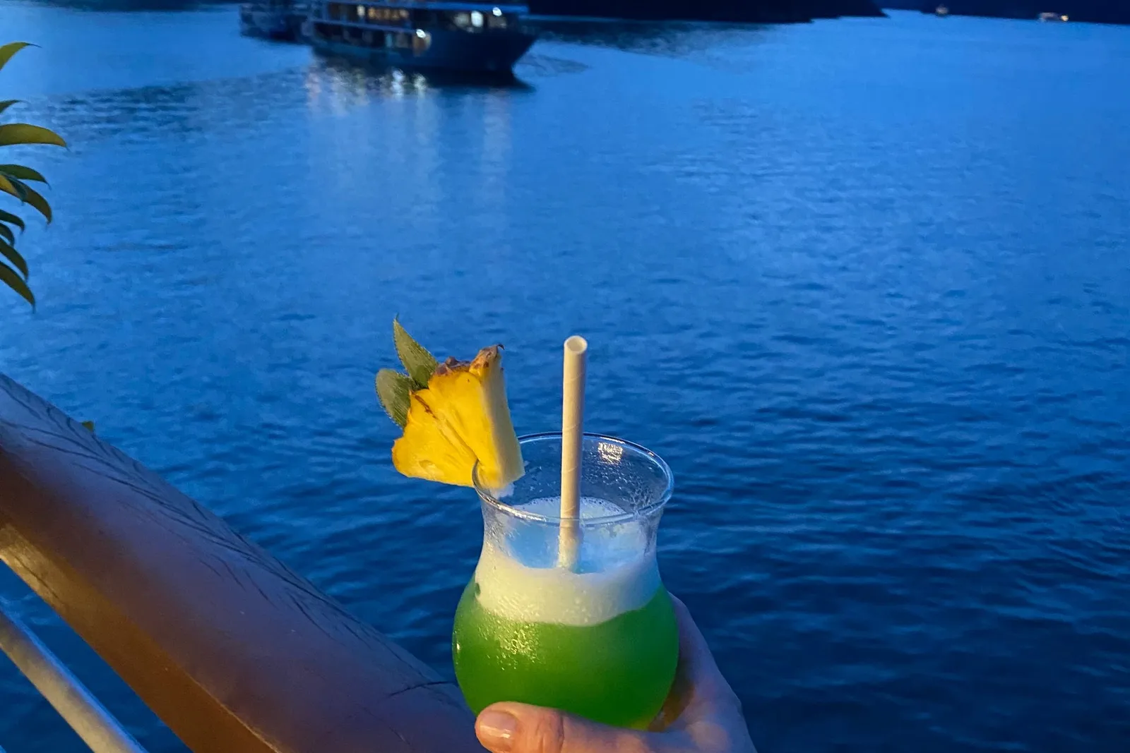 A tattooed hand holds a green cocktail with a pineapple wedge and paper straw against a wooden boat railing, with a lit cruise boat anchored in a bay of dark limestone karsts at dusk.