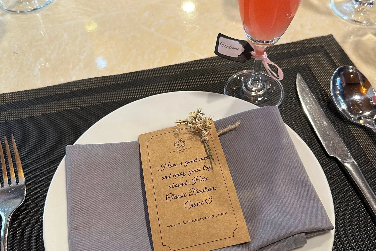 A dining table place setting with a grey folded napkin on a white plate topped with a kraft card reading 'Have a good meal and enjoy your trip aboard Hera Classic Boutique Cruise', beside a pink drink in a flute glass with a 'Welcome' tag.